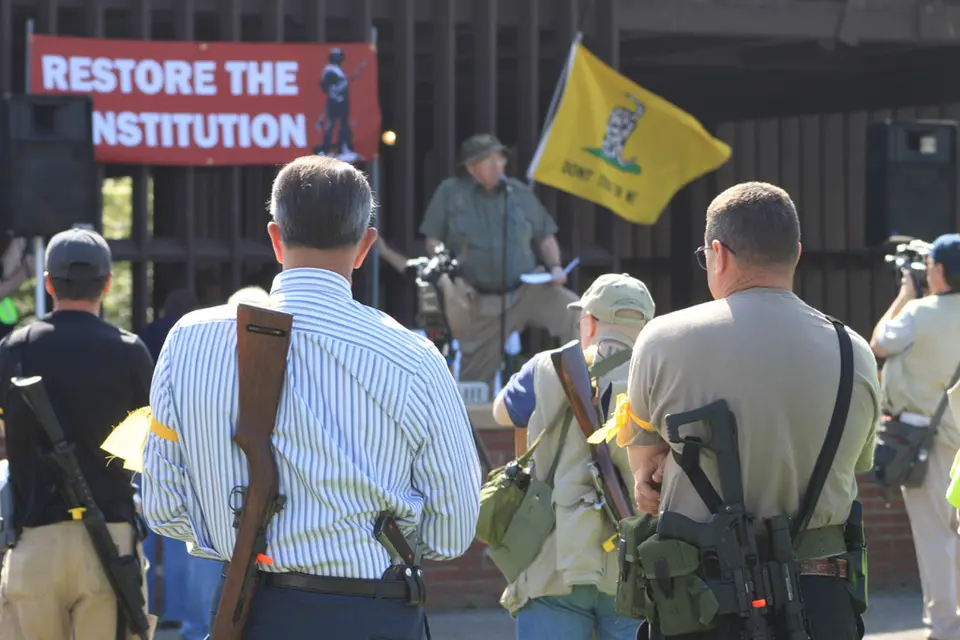 Men with guns stand outside during a tea party rallly in 2016 with a restore the consitution banner in the background